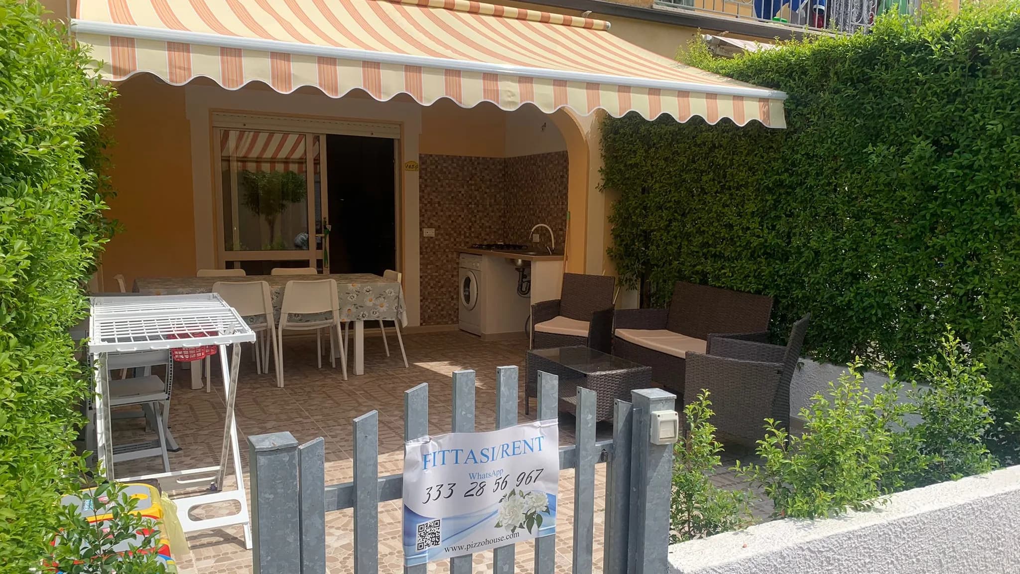 Veranda della casa con tavolo, cucina esterna e salottino, vista dal cancello d'ingresso